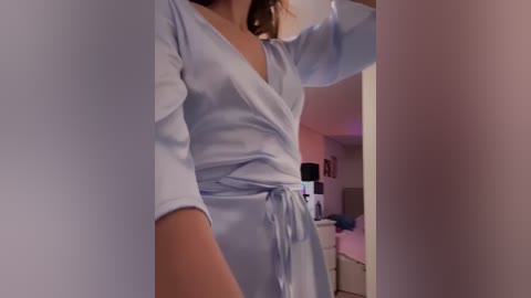 Media: Video of a woman in a light blue satin robe, standing in a dimly lit bedroom with a bed, dresser, and wall art visible in the background.