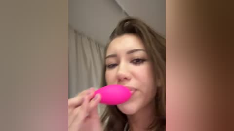 Media: Video of a light-skinned woman with brown hair, wearing black eyeliner, using a bright pink sex toy, in a dimly lit bathroom.