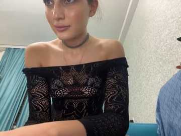 Media: A video of a young woman with light skin and dark hair, wearing a black lace top and a choker necklace, in a room with teal curtains and textured wallpaper.