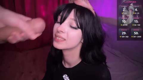 Media: Video of a young Asian woman with long black hair, smiling, being touched on the head by a large, erect penis. Background includes a purple-lit room and a digital screen showing stats.