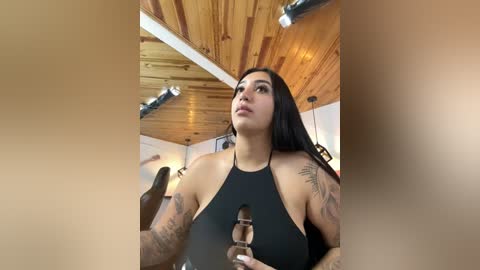 Media: Video of a tattooed Latina woman with long black hair, wearing a black halter top, in a rustic wooden room with exposed beams.