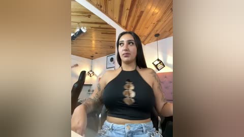 Media: Video of a Latina woman with long dark hair, medium skin tone, and tattoos on her arms, wearing a black halter top and high-waisted jeans, sitting in a cozy, wooden-paneled room with a plush chair and modern lighting.