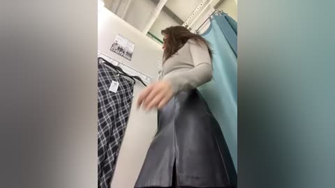 Media: Video of a woman in a beige sweater and black skirt, browsing clothes in a store, with a teal garment hanging nearby.