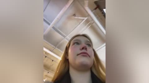 Media: Video of a young woman with light skin and long blonde hair, standing indoors with an industrial ceiling visible above. She looks upward with a contemplative expression.