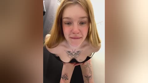 Media: Video of a young, fair-skinned, blonde woman with tattoos on her chest, arms, and neck, leaning forward, wearing a black bra.