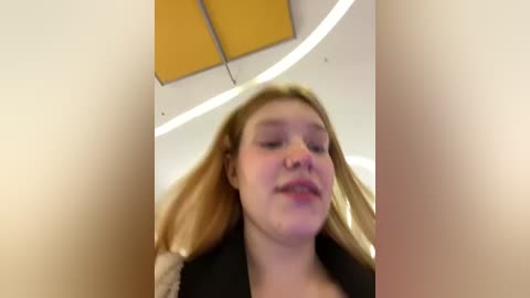 Media: Video of a young, fair-skinned woman with long blonde hair, wearing a black coat, looking upward with a neutral expression. Background shows a modern, curved ceiling with yellow accents.