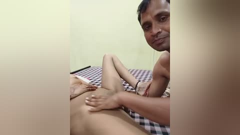 Media: A video of a South Asian man with short black hair, medium build, lying on a bed with a checkered blanket, wearing only a black wristband, touching the genitals of another person.