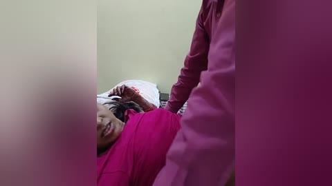 Media: Video of a young woman with medium skin tone, lying on a bed in a pink shirt, smiling, with a person in a pink shirt standing over her, partially blurred.