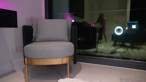 Media: Video of a modern, grey and white checkered armchair with gold legs and cushions, positioned in a sleek, minimalist living room with large glass windows reflecting a woman dancing.