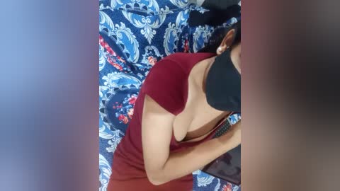 Media: A video of a woman in a red dress, partially revealing cleavage, lying on a blue patterned bedspread. Her head is partially covered by a black mask.