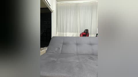 Media: Video of a woman in a red dress adjusting a gray, tufted sofa against a white curtain backdrop.