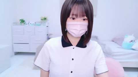 Media: Video of an Asian woman with short brown hair, wearing a pink face mask and white polo shirt, sitting in a minimalistic white bedroom with a dresser and bed.