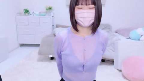 Media: Video of an Asian woman with straight brown hair, wearing a light purple sheer top, face mask, and black pants, standing in a modern, white minimalist bedroom with a white dresser, bed, and pink and blue balloons.