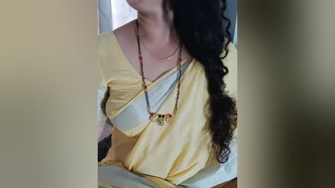 Media: Video of a woman with long, curly black hair, wearing a yellow saree with a white border, adorned with a red and black beaded necklace. The background is blurred, focusing on her attire and jewelry.