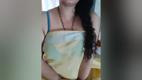 Media: Video of a woman with medium skin tone, wearing a yellow sari with a blue border and a red blouse. She has long, curly black hair. Background shows a blurred, neutral-toned room with white walls.