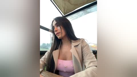 Media: Video of a young woman with long dark hair, wearing a beige blazer over a pink floral top, seated in a modern, sunlit room with large windows.