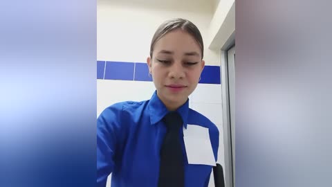 Media: Video of a young woman with light skin and brown hair, wearing a blue and black uniform, standing in a bathroom with white and blue tiled walls.