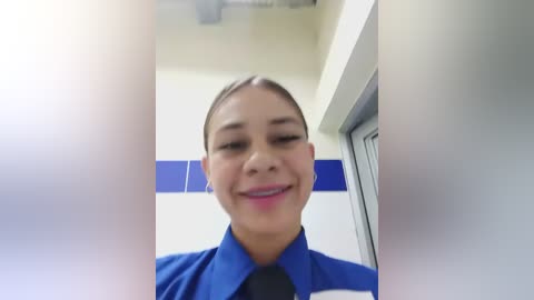 Media: Video of a smiling Latina woman with dark hair in a bun, wearing a blue and white uniform, standing in a brightly lit office hallway with blue accents.