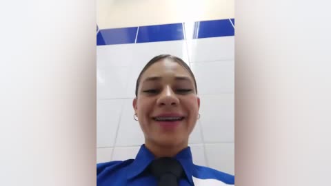 Media: Video of a smiling, light-skinned woman with dark hair, wearing a blue uniform shirt and black tie, standing in a white-tiled bathroom with blue accents.