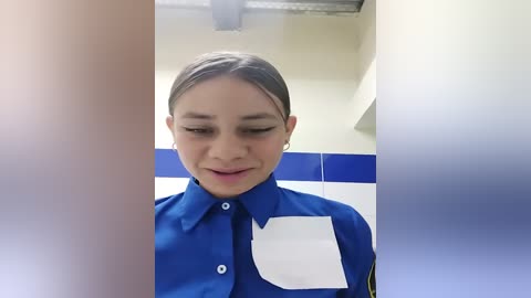 Media: Video of a young woman with light skin, brown hair tied back, wearing a blue uniform with white patches and a badge, smiling in a bright, tiled room with blue accents.