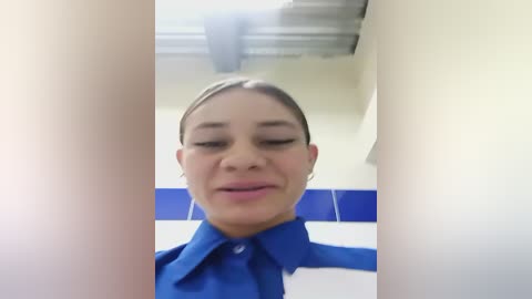 Media: Video of a young woman with light skin, brown hair, and brown eyes, wearing a blue shirt and tie, standing in a bathroom with white tiles and blue trim. She has a slight smile.