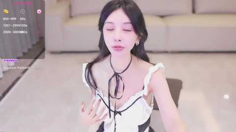 Media: Video of a young Asian woman with long black hair, wearing a revealing black and white maid outfit with a large bow. She kneels on a light-colored floor in a modern living room.