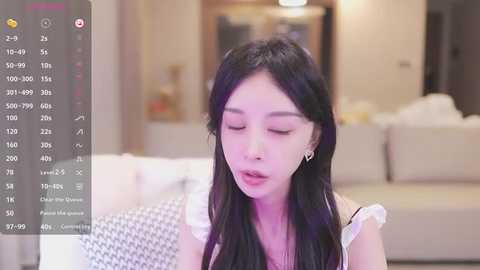 Media: Video of a young Asian woman with long black hair, wearing a white off-shoulder top, sitting on a couch in a modern living room. The background shows a blurred sofa and neutral decor.