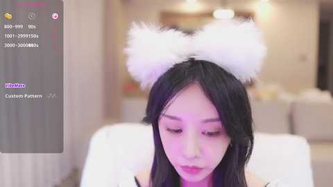 Media: Video of a young Asian woman with long black hair, wearing white fluffy cat ears, in a soft-lit living room.