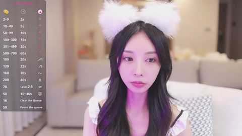 Media: Video of an East Asian woman with long black hair, wearing a white fluffy cat headband and a white lace top, indoors with a blurred background.