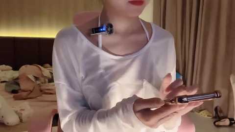 Media: Video of a person with fair skin wearing a white long-sleeve shirt, holding a vaping device with a blue light, in a dimly lit bedroom with a bed and stuffed toys.