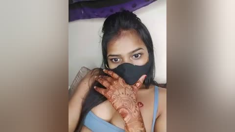 Media: Video of a South Asian woman with medium skin tone, black hair, wearing a blue bra, black mask, and intricate henna design on her hands. Background features a purple curtain and plain wall.