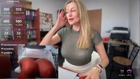 Media: Video of a blonde woman in a green sweater and red tights, holding a phone, in a cluttered room with a bookshelf, desk, and monitor.