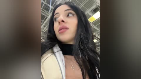 Media: Video of a young woman with long, black hair and fair skin, wearing a black top and beige jacket, standing in a modern, industrial building with a high ceiling and visible beams.