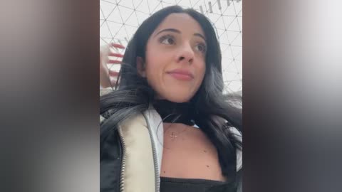 Media: A video of a young woman with long black hair and light skin, wearing a black jacket and a black top, standing near a chain-link fence.