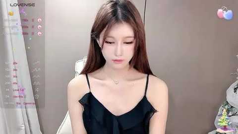 Media: Video of a young East Asian woman with long brown hair, wearing a black spaghetti-strap top, sitting indoors, looking downward. Background includes a gray wardrobe and a white chair.