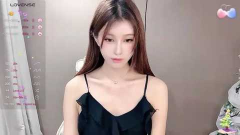 Media: Video of a young Asian woman with fair skin, long brown hair, wearing a black camisole, sitting in a modern, minimalist room with beige walls and a calendar on the left.