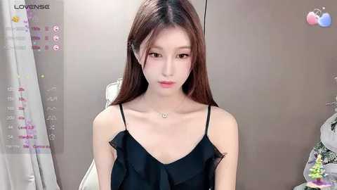 Media: A video of an East Asian woman with long brown hair, wearing a black sleeveless top, in a modern, minimalist room with a gray wall and white curtain.