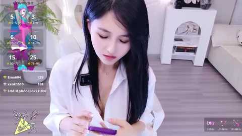 Media: A video of an East Asian woman with long black hair, wearing a white shirt, sitting on a white sofa, looking at a smartphone, in a modern living room.