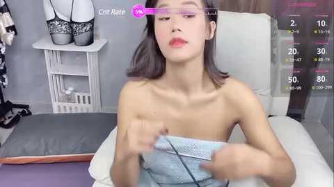 Media: A video shows a topless Asian woman with fair skin, brown hair, and light makeup, wearing a blue towel. She's in a modern, well-lit room with grey walls and a white bench.