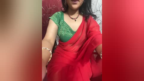 Media: Video of a South Asian woman in a red saree with green blouse, wearing a necklace and bracelets, sitting on a red-patterned cushioned chair against a white wall, smiling.