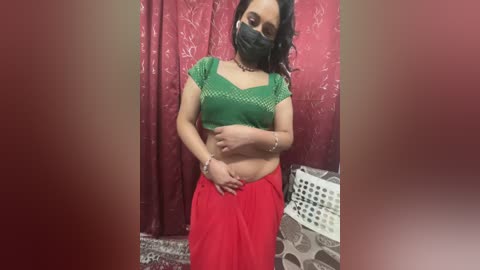 Media: A video of a South Asian woman with medium skin tone, wearing a green crop top, red skirt, black face mask, and a silver bracelet, standing in a room with red curtains and a white laundry basket.