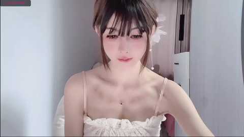 Media: Video of an Asian woman with fair skin, wearing a white lace dress, sitting in a room with white walls and a wooden door. She has straight black hair and a neutral expression.