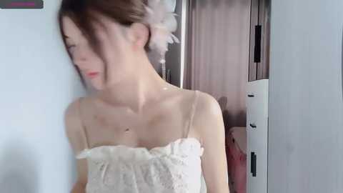 Media: Video of a slender, light-skinned woman with brown hair styled in an updo with a white flower, wearing a lace, off-shoulder wedding dress. She stands in a hallway, partially obscured by a door.