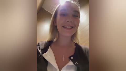 Media: A video of a smiling, fair-skinned young woman with straight, shoulder-length blonde hair, wearing a black blazer over a white shirt, taken indoors with warm lighting.