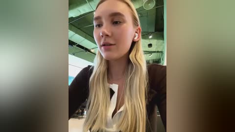 Media: Video of a young woman with long, straight blonde hair, wearing a white blouse and a black cardigan, standing indoors in a modern, industrial-style room with exposed pipes and green lighting.