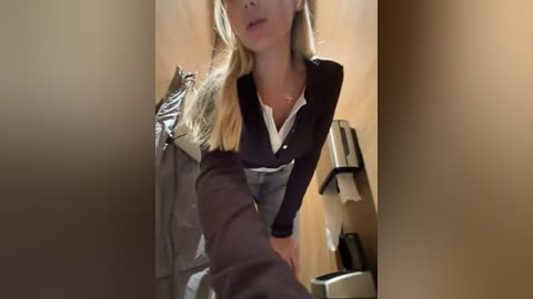 Media: A video captures a blonde woman in a dark blazer over a white shirt, holding a tissue, in a beige bathroom with a metallic trash can and toilet paper roll holder.