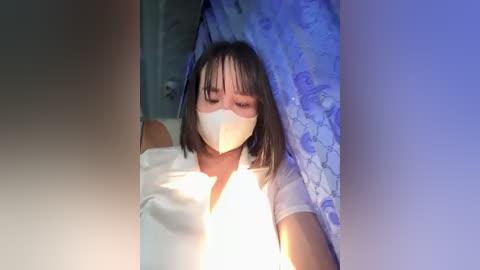 Media: A video of a young woman with straight brown hair, wearing a white face mask, white shirt, and blue-patterned curtain. She appears to be in a bathroom, seated on a toilet, with a bright light illuminating her from the left.