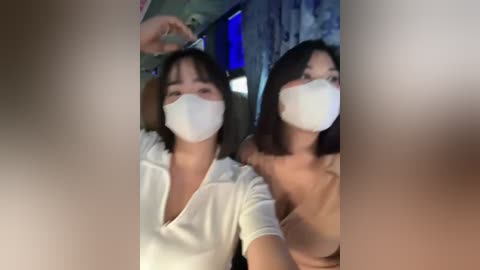 Media: Video of two East Asian women with straight black hair, wearing white face masks, white tops, and sitting close together. Background shows a dimly lit room with blue and green hues.