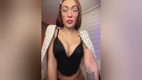 Media: Video of a young, light-skinned woman with glasses and brown hair, wearing a black lace bra and a white jacket, displaying tattoos on her arm, standing indoors with white blinds in the background.