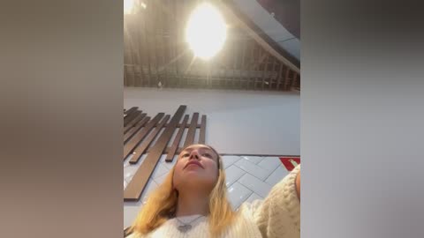 Media: Video of a young woman with long blonde hair, wearing a beige sweater, standing in a bright, modern room with a large ceiling light and metallic shelving unit in the background.